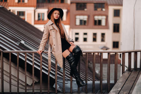 A stylish young woman in a beige coat and black hat sits on a rooftop in the city center. Women's street fashion. Autumn clothing.Urban style.の写真素材