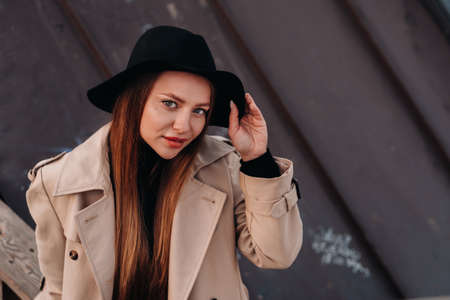 A stylish young woman in a beige coat and black hat sits on a rooftop in the city center. Women's street fashion. Autumn clothing.Urban style.の写真素材