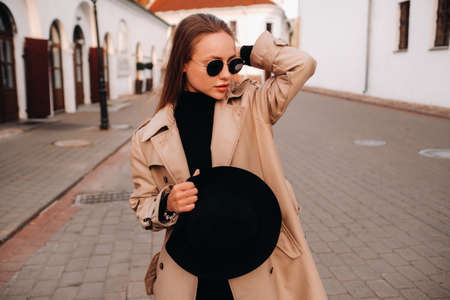 Stylish young woman in a beige coat and a black hat in his hands and glasses on a city street....