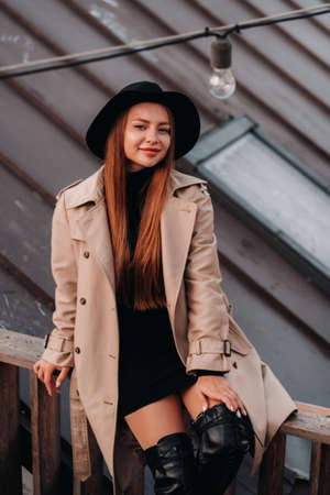 A stylish young woman in a beige coat and black hat sits on a rooftop in the city center. Women's street fashion. Autumn clothing.Urban style.の写真素材