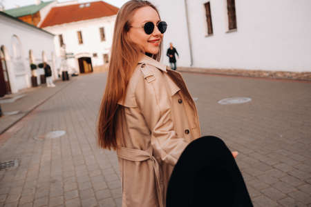 Stylish young woman in a beige coat and a black hat in his hands and glasses on a city street. Women's street fashion. Autumn clothing.Urban styleの写真素材