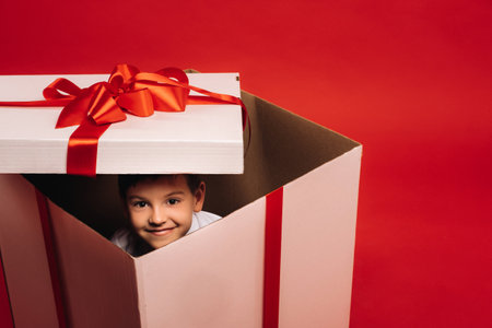 A little boy sits in a Christmas present and looks out of it on a red background.の写真素材