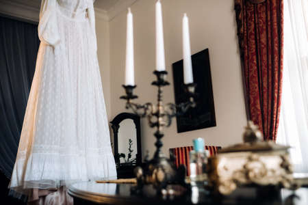 Vintage wedding dress hanging on a wooden hanger and a candle holder standing on a table in the castle.の写真素材