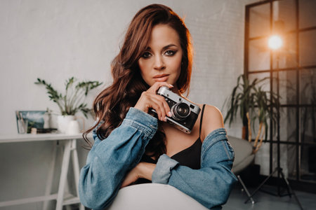 a brunette with long hair poses in a studio sitting on a chair with a camera in her hands.の写真素材