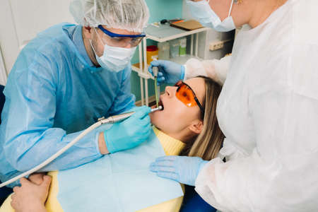 A male dentist with dental tools drills the teeth of a patient with an assistant. The concept of...