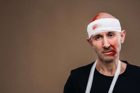 A battered man with a bandaged head and a cast on his arm stands on a gray background.の写真素材