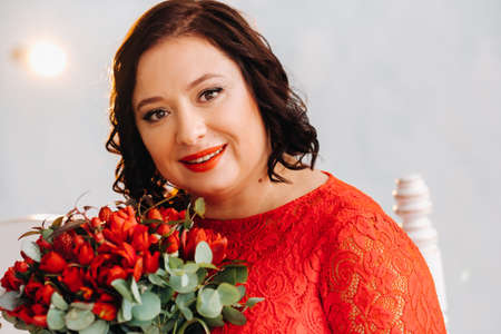 a woman in a red dress sits on a sofa and holds a bouquet of red roses and strawberries in the interiorの写真素材