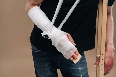 A battered man with a bandaged head and a cast on his arm stands on crutches on a gray background.の写真素材