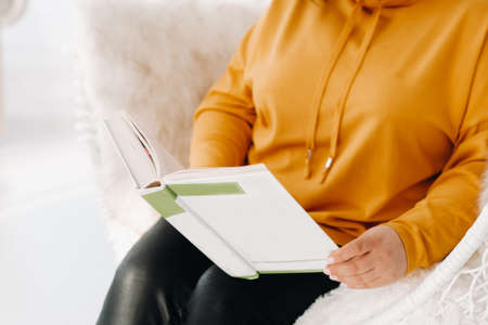 a woman in an orange hoodie sits in an unusual chair with a book in her hands.の写真素材