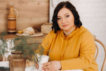a woman in an orange hoodie is sitting at a table in the kitchen drinking coffee.の写真素材