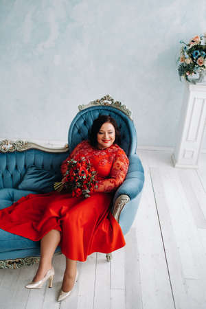 a woman in a red dress sits on a sofa and holds a bouquet of red roses and strawberries in the interiorの写真素材