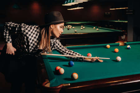 A girl in a hat in a billiard club with a cue in her hands hits a ball.Playing pool.の写真素材