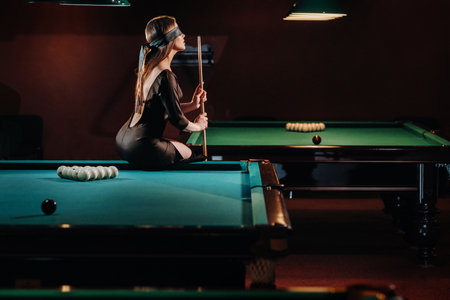 A girl with a blindfold and a cue in her hands is sitting on a table in a billiard club. Russian...