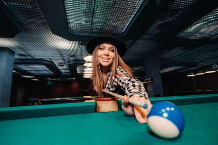 A girl in a hat in a billiard club with a cue in her hands hits a ball.Playing poolの写真素材