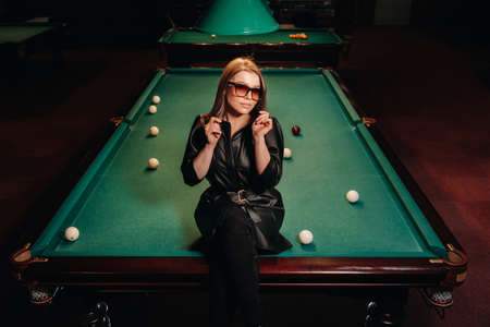 A girl with glasses sits on a pool table in a club.Russian billiards.