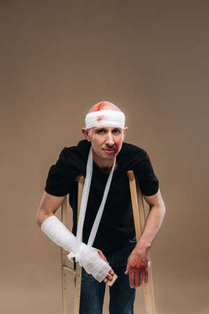 A battered man with a bandaged head and a cast on his arm stands on crutches on a gray background.の写真素材