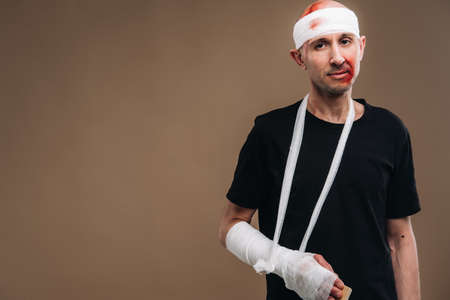 A battered man with a bandaged head and a cast on his arm stands on crutches on a gray background.の写真素材