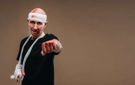 A battered man with a bandaged head and a cast on his arm stands on a gray background.の写真素材