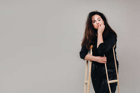 A woman beaten in black clothes with crutches in her hands on a gray background.の写真素材