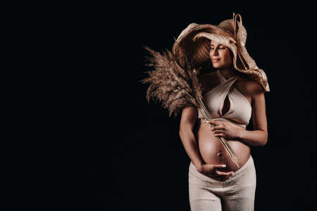 pregnant woman in a straw hat with beige clothes with a bouquet in her hands in the studio on a black background.の写真素材
