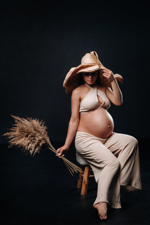 pregnant woman in a straw hat with beige clothes with a bouquet in her hands in the studio on a black background.の写真素材
