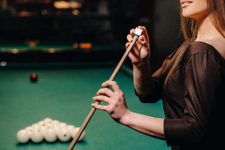 A girl in a dress stands with a cue in her hands and cleans it with chalk in a billiard club with balls in her hands.Playing billiardsの写真素材