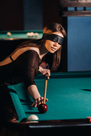 A girl with a blindfold and a cue in her hands in a billiard club.Russian billiards