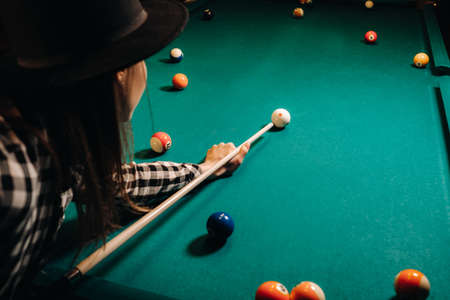 A girl in a hat in a billiard club with a cue in her hands hits a ball.Playing poolの写真素材