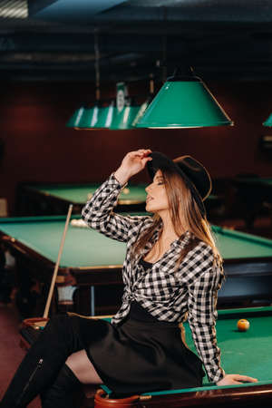 A girl in a hat in a billiard club sits on a billiard table.Playing poolの写真素材