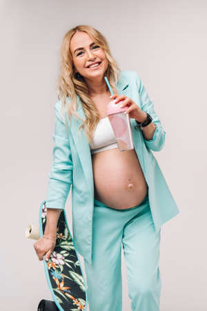 pregnant girl in turquoise clothes with a glass of juice and a skateboard in her hands on a blue background.の写真素材
