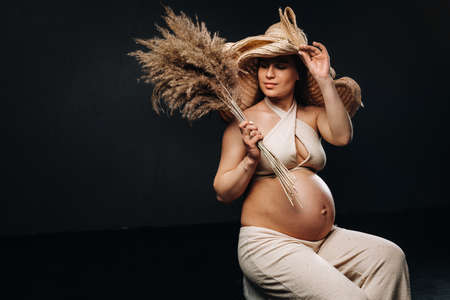 pregnant woman in a straw hat with beige clothes with a bouquet in her hands in the studio on a black background.の写真素材
