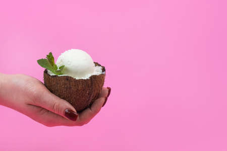 Vanilla ice cream balls in a fresh coconut half, decorated with mint leaves in your hand on a pink background.の写真素材