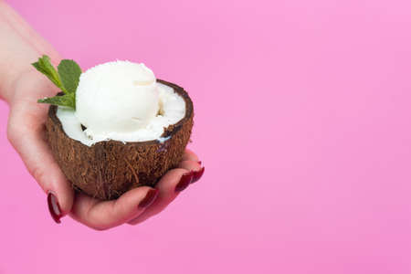 Vanilla ice cream balls in a fresh coconut half, decorated with mint leaves in your hand on a pink background.の写真素材