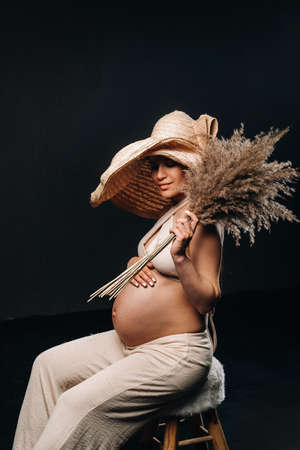 pregnant woman in a straw hat with beige clothes with a bouquet in her hands in the studio on a black background.の写真素材