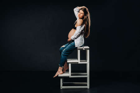 pregnant woman sitting on a ladder chair in a studio on a black background.