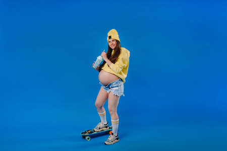 a pregnant girl in yellow clothes with a glass of juice rides a skateboard on a blue background