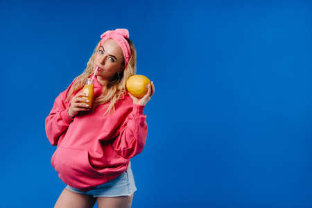 pregnant girl in pink clothes with a bottle of juice and a melon on a blue backgroundの写真素材