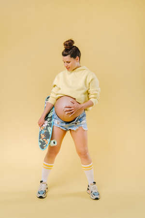 Pregnant girl in a yellow jacket with a skateboard in her hands on a yellow background.の写真素材