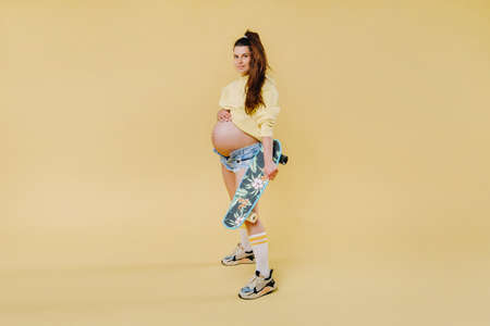 Pregnant girl in a yellow jacket with a skateboard in her hands on a yellow background.