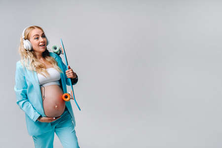 A pregnant girl in a turquoise suit with a skateboard in her hands and headphones stands on a gray...