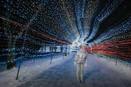 a little girl in a silver jacket in winter with evening burning lights on a Christmas decorated street.の写真素材