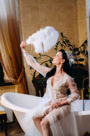 the bride, dressed in a boudoir transparent dress and underwear, sits on a vintage bathtub with a...