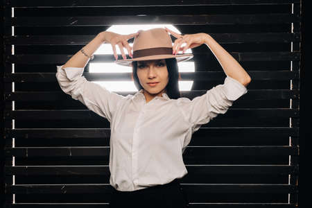 Elegant brunette girl in a white shirt and beige hat on a dark background.