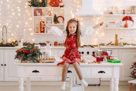 A happy little girl in the Christmas kitchen is sitting on the table with cookies in her hands.の写真素材