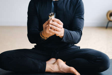 A male athlete in black sportswear sits in the lotus position in his hands with an incense candle in...