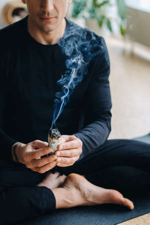 A male athlete in black sportswear sits in the lotus position in his hands with an incense candle in the gym.の写真素材