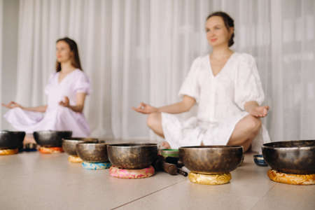 Two women are sitting with Tibetan bowls in the lotus position before a yoga class in the gymの写真素材