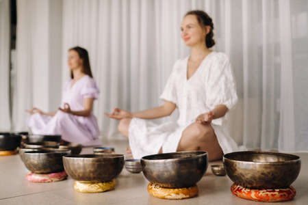 Two women are sitting with Tibetan bowls in the lotus position before a yoga class in the gym.の写真素材