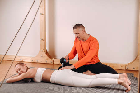 A male masseur massages a girl's body with a percussion massager to prepare for a stretching...
