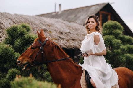 A woman in a white sundress riding a horse near a farm.の写真素材